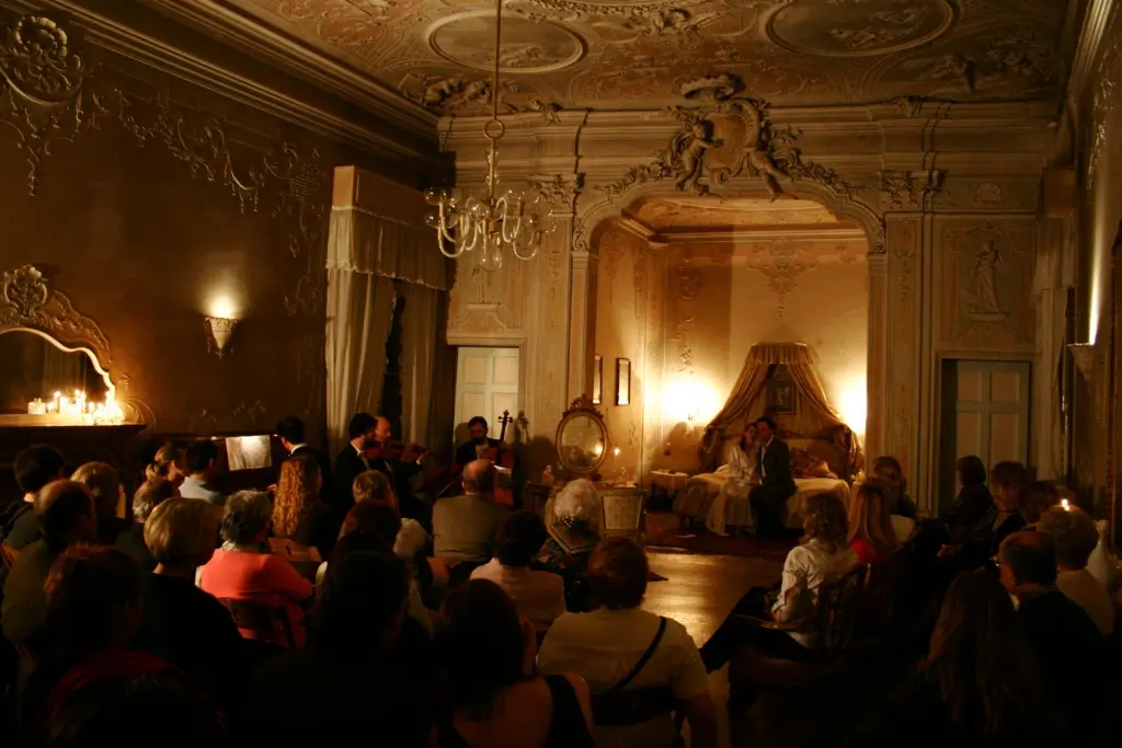 An audience seated on chairs in a candlelit palace salon, with two singers on a bed in an alcove and musicians playing to the left
